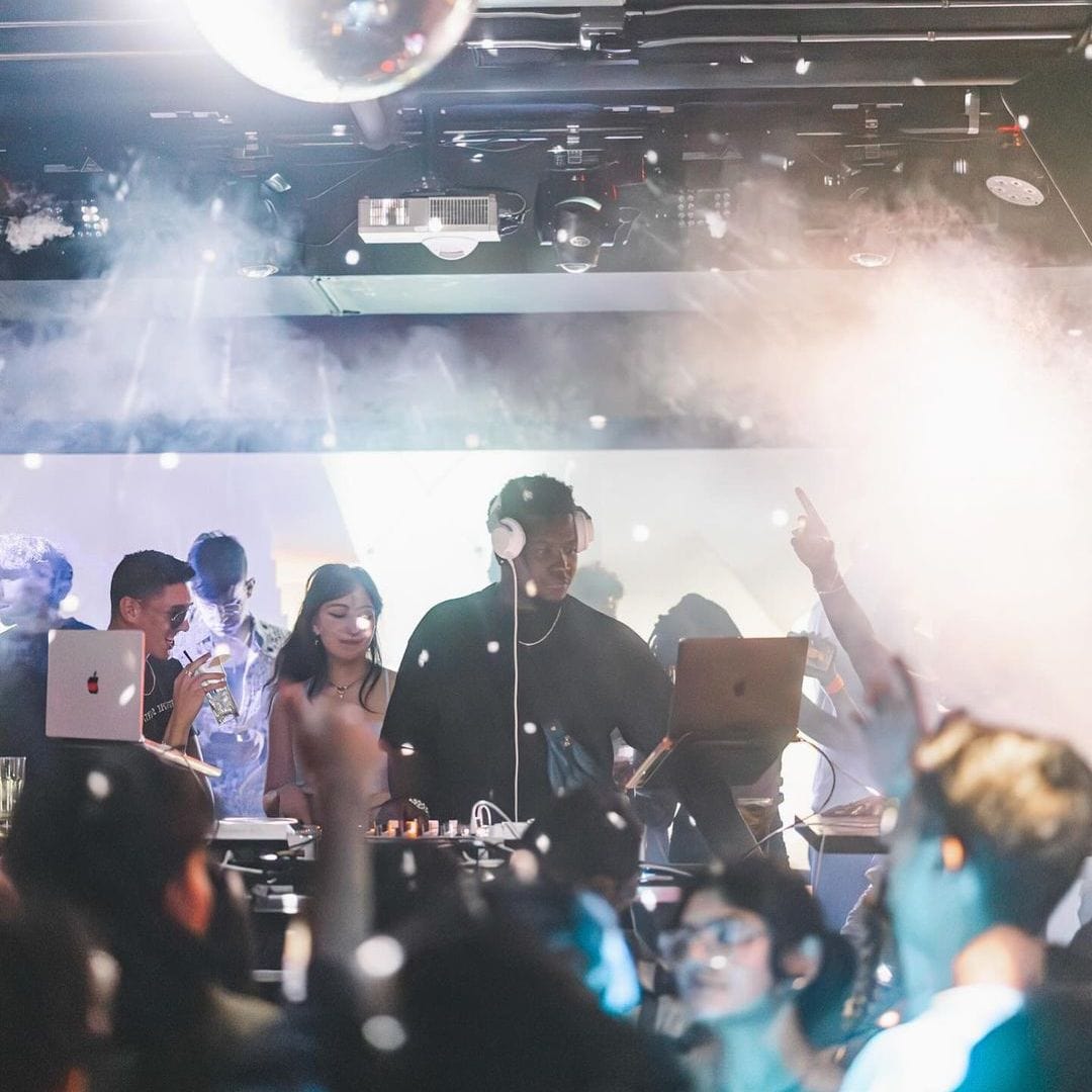 Producer wearing white headphones performing as a DJ in a nightclub on stage with people dancing and smoke effects.