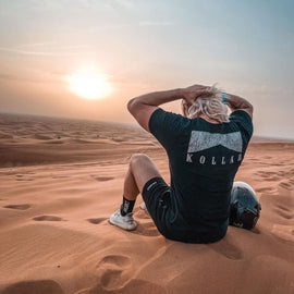 A blonde-haired man wearing a black Kollar t-shirt sits on the sand in a vast desert, gazing at the sunset with his hands on his head. A motorcycle helmet rests beside him, symbolizing adventure and exploration